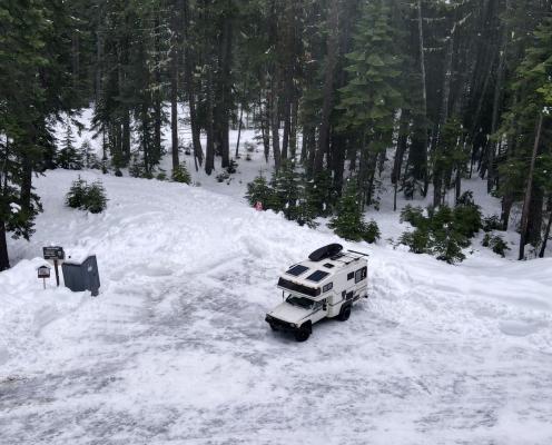 Parking area covered by bumpy ice and snow surrouned by thick snow-covered ground and pine trees. A truck is parked in the middle and a porta-potty sits on the left edge of the lot.