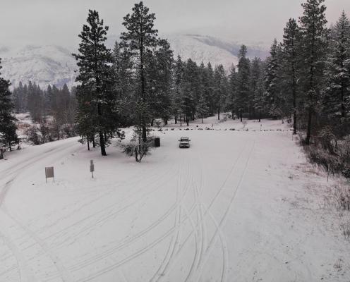 A snow covered parking lot surrounded by pine trees with snow-dusted mountains in the background.