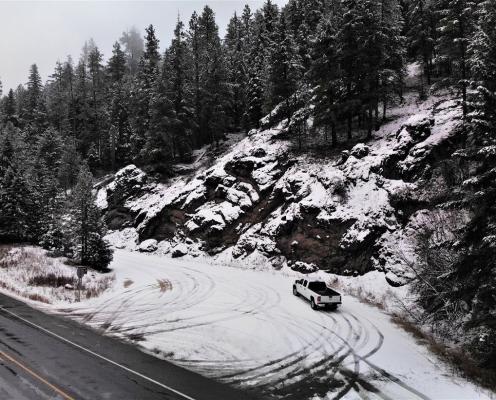 A snow-covered turn-off from a plowed highway with a wall of sloping rocks and trees on the right side dusted in snow.