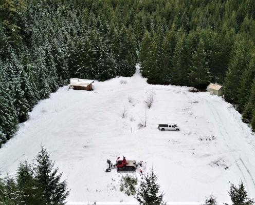 Overhead view of a square parking area covered by thick snow and bordered by a thick pine tree forest. A warming hut sits in the upper left corner and pit toilet hut sits on the upper right corner.
