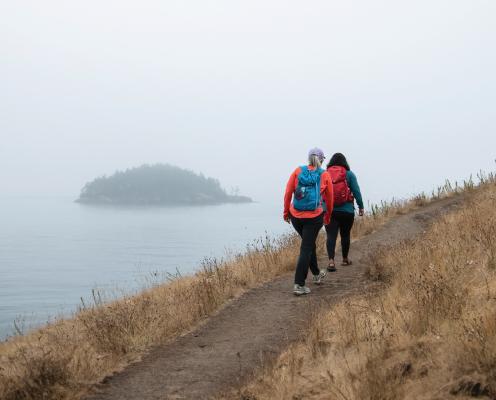 Two female hikers in orange and blue hike a bluff trail above the water with an island in the background.