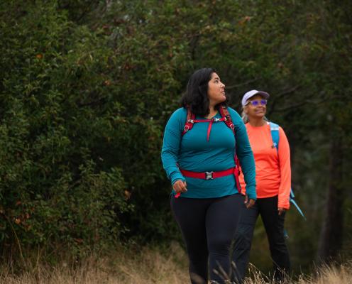 Two women, one in blue and one in orange, hike in the forest wearing backpacks.