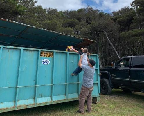 A man lifts a boy to throw trash into a blue dumpster. 