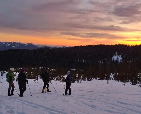 Four snowshoers stand in front of a partly cloudy winter sunrise.