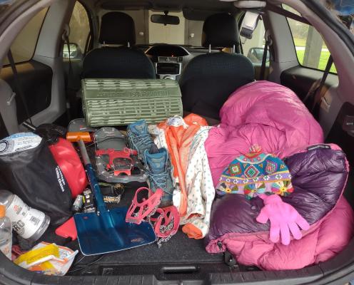 An open car trunk with winter survival gear: tire chains, gas can, water, food, snowshoes and microspikes, shovel and mats to help get a car unstuck in the snow, quilt, reflective clothing, warm layers.