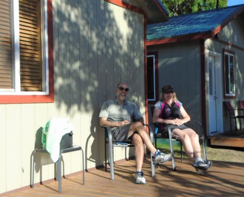 A middle-aged male and female sit on chairs on the deck of a wood cabin in the sun.
