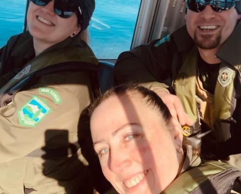 Three young people wearing Parks uniforms sit at the helm of a Parks boat on the water. The photo is taken as a group selfie.