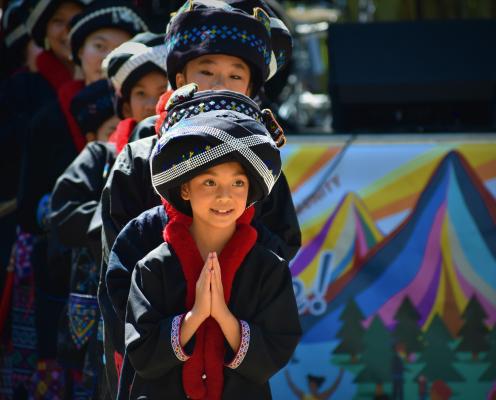 A group of young Mien dancers dress in traditional clothing. The girl in the front smiles and holds up her hands in praying gesture. 