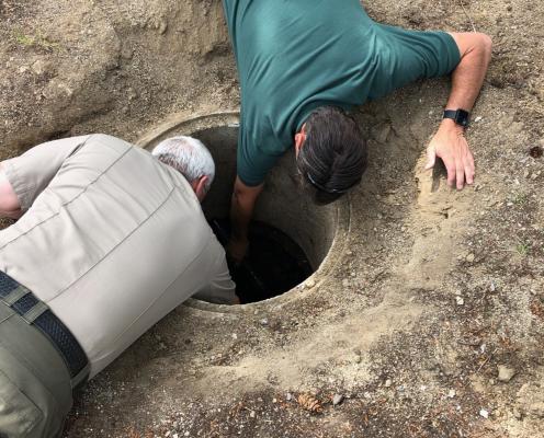 Two men, one in a park aide shirt and one in a ranger uniform, look down a hole in the ground to examine a septic system.