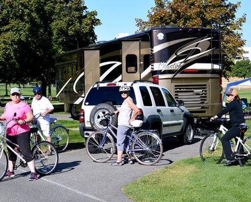 A group of cyclists pose in a campground in front of an RV and car.