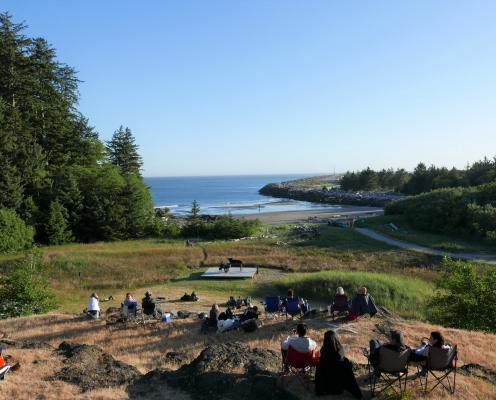 People sit on a hill overlooking a beach, watching a concert.