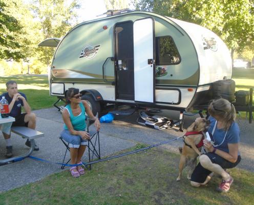 A woman pets a couple's dog as they sit outside a tiny trailer.