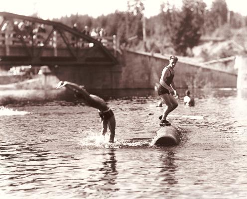Two young men stand on logs in a river with a bridge in the background in this historic sepia toned photo.