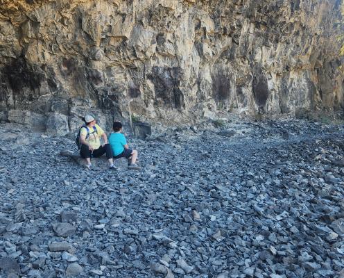 A  woman and a boy sitting and relaxing as they just did the Lake Lenore Caves hike to one of the caves. The cave is super rocky and shaded away from the sun. The cave is very shallow.