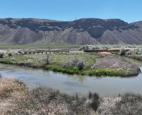 Large basalt hill and blue sky overlooking two trestles, one over water and one over land connecting a straight trail. 