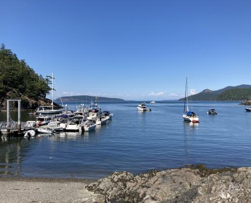 Course, grey sand beach with grey rocks on the right meet the blue water in the foreground. On the left are numerous motor boats tied to a long dock, the edge of a treed, rocky hillside sit behind them. Several motor and sail boats sit in the bay looking towards the open water. A hand full of treed islands in front of cloudy horizon with a blue sky in the background.