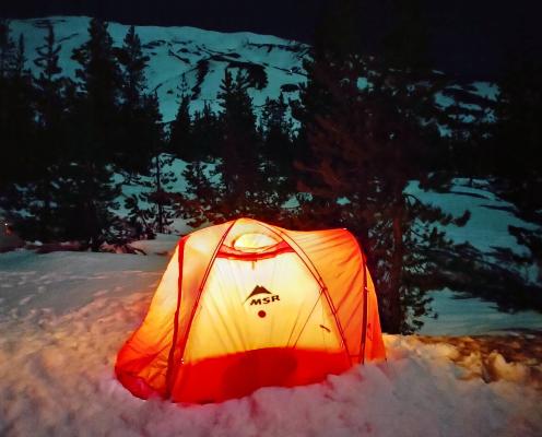 An orange tent illuminated from inside below a dark snowy mountain.
