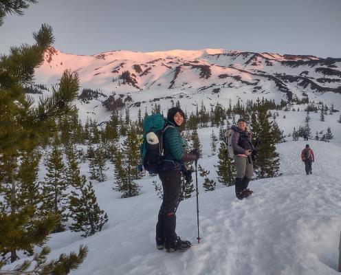 Two women snowshoe at sunrise below a snowy mountain.
