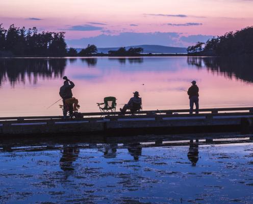 Three fishermen on a pier in the sunset. Two are standing, one is sitting.