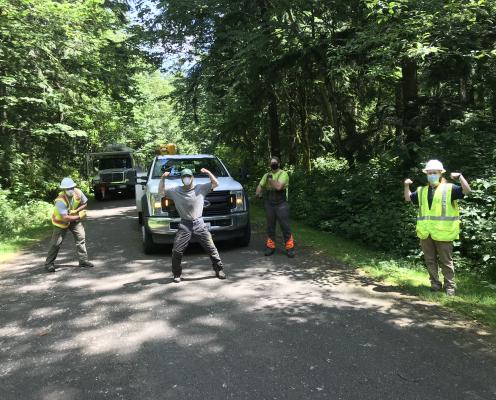 Three men in safety vests pose in front of tree cutting equipment.