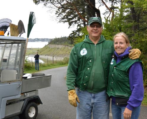 A couple stands next to a golf cart with gardening equipment in front of a beach.