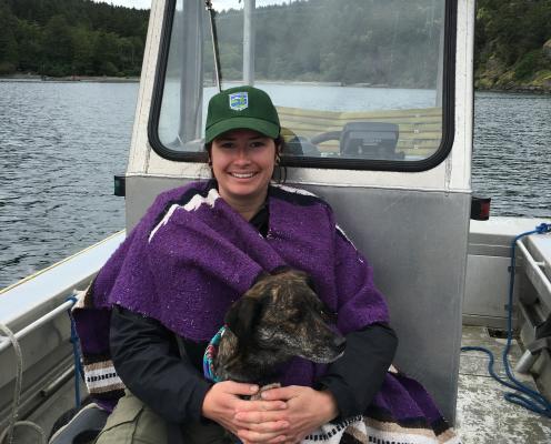 A woman in a Parks hat sits in an aluminum boat with a blanket and a brown dog.