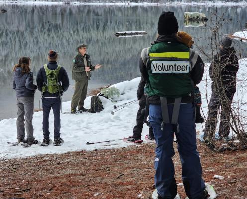 A ranger gives a talk by a snowy lake, as a volunteer and visitors listen.