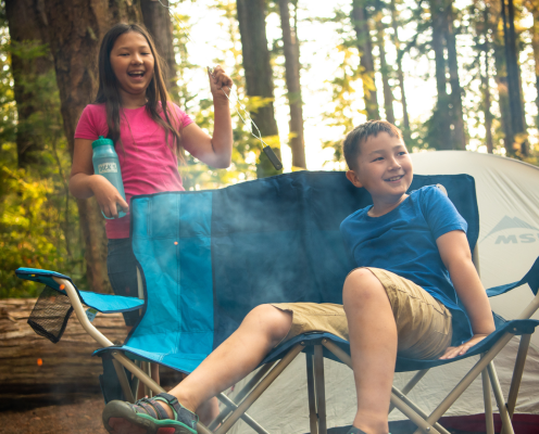 Boy sitting in a campchair with a girl behind him holding a roasting stick with a tent in the background