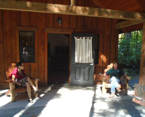 Two women, one in a State Parks uniform, sit under a covered porch in front of a wood cabin talking.