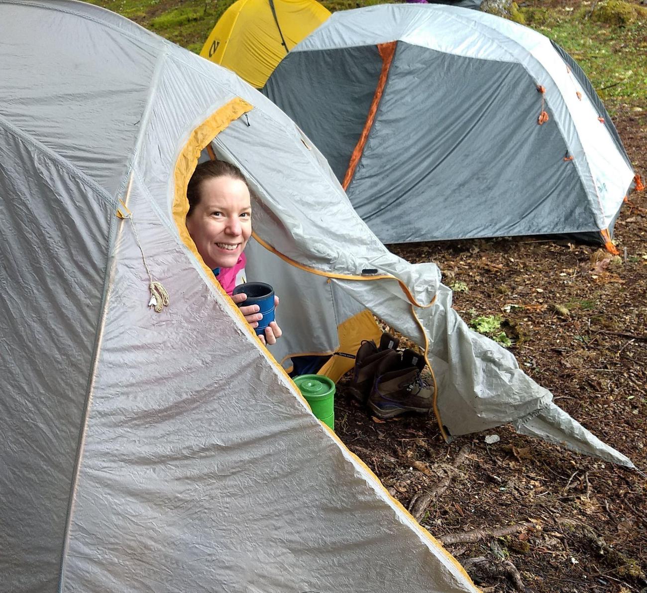 A person sticks their head out of their tent door with a beverage mug. A 2nd tent in the background