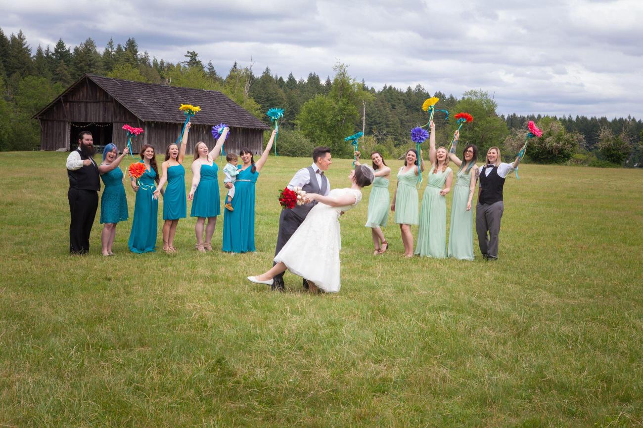 A newlywed couple poses in a field with their wedding party in blue and green in the background.