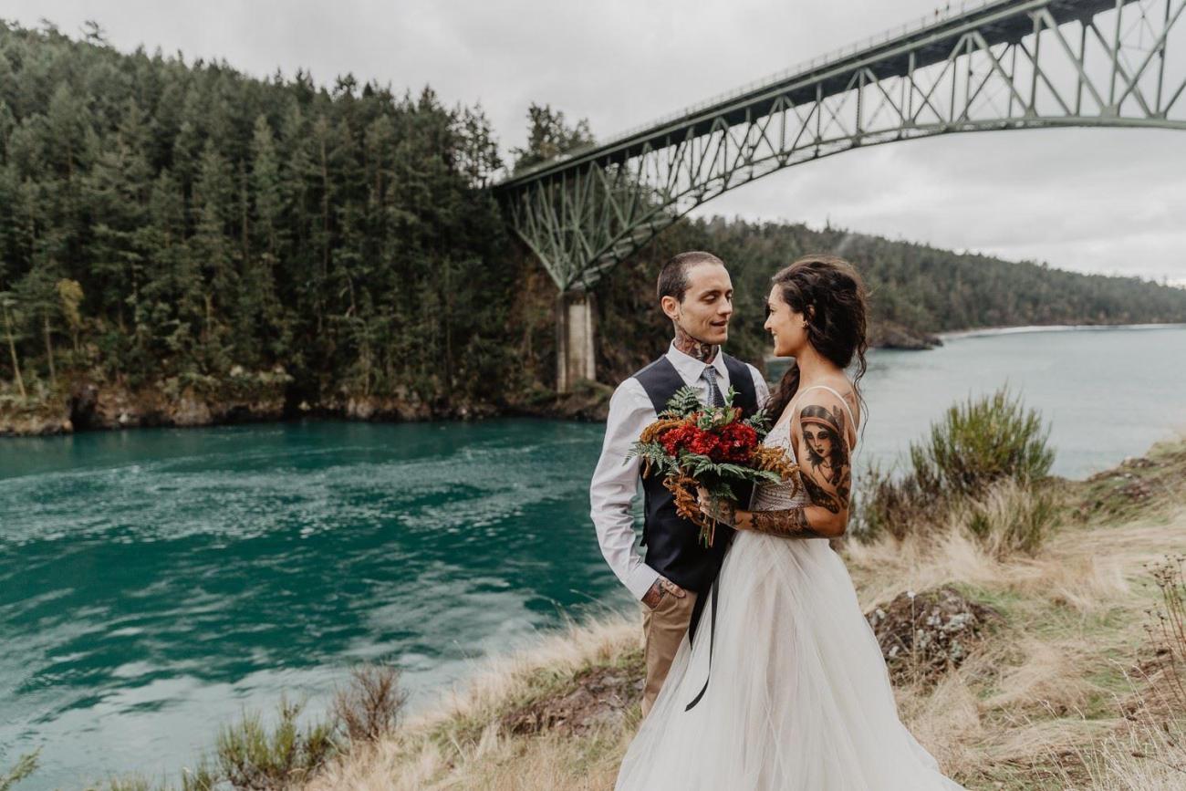 A couple stands on a cliff in front of a bridge with water below.