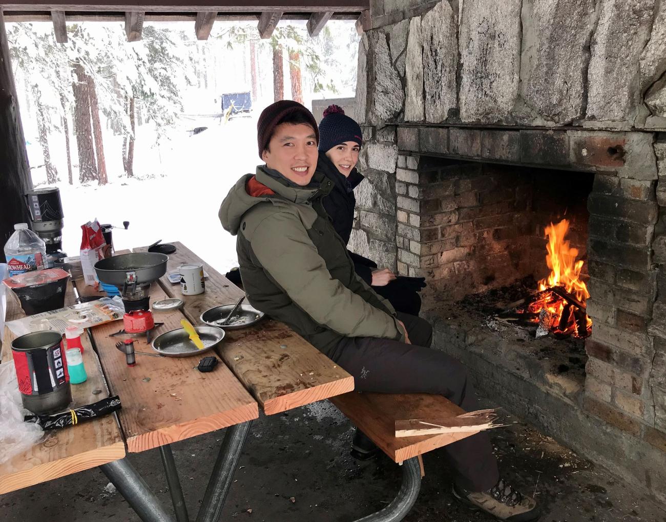 A couple sits on a picnic table bench in front ouf a stone outdoor fireplace