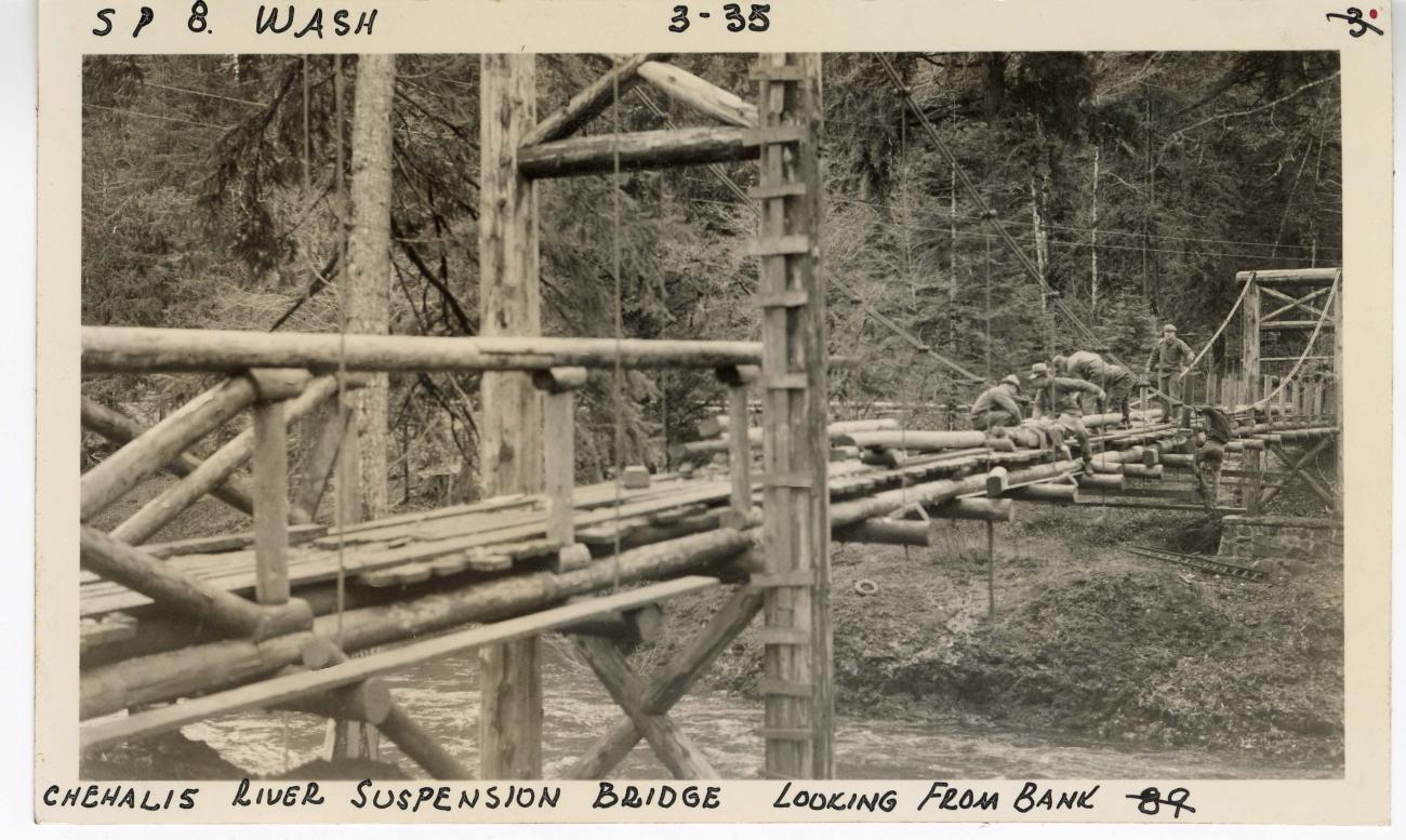 black and white historic photo of workers building timber suspension bridge over a river