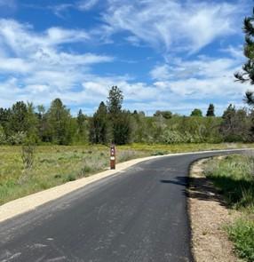 The newly paved Centennial Trail with fresh gravel along one shoulder. Green grass, a line of trees, and blue sky are in the background.