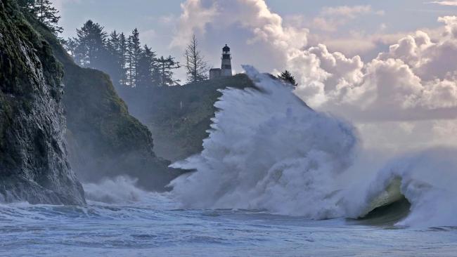 High king tide wave crashes against rocky cliffside with a lighthouse