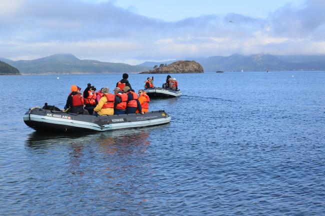Boaters in a dinghy with red and orange life vests. One is waving.