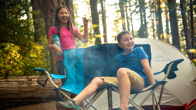 Boy sitting in a campchair with a girl behind him holding a roasting stick with a tent in the background