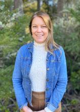 Heather Saunders, Washington State Parks Development Director stands in front of evergreens wearing a blue shirt and brown pants.