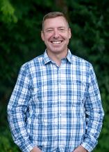 Washington State Parks Operations Director Jon Crimmins poses in front of evergreens smiling and wearing a blue plaid shirt. 