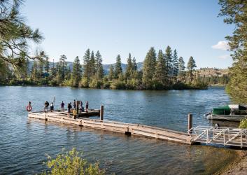 A group of people stand at the end of a dock fishing into the lake with tall trees and bushes along the far shore.