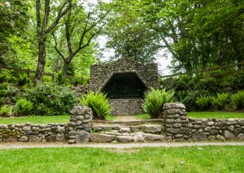 The grotto is made up of hundreds of stones of varying colors surrounded by multiple bushes and lush green trees. There is a path leading up to the grotto with a few steps. 
