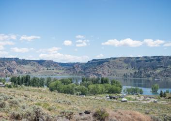 View of campground with green trees and the lake behind, reflecting the basalt rock hillside in the background