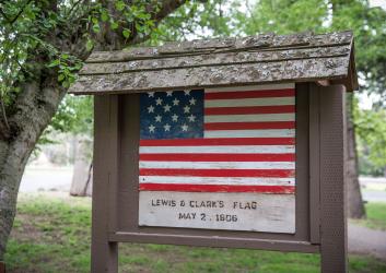 Brown roofed sign with a painted replica of Lewis and Clark's flag from 1806 with 15 stars on a blue background and 15 red and white stripes