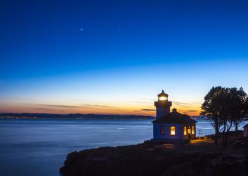 An illuminated lighthouse sits on a rocky cliff overlooking water as the sun sets behind and the sky grows dark. A star shines bright in the sky.