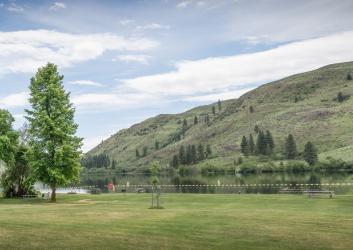 Grassy lawn scattered with full, green trees and picnic tables leading to the water's edge with a buoy swim line calmly sitting on the water.