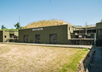 A cement battery building sits with brown grass in front and on top. Doors are boarded shut with stair ways throughout the structure.