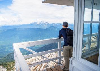 A hiker takes a break while enjoying the view of forested mountains and a snowcapped mountain with a cloudy sky from the lookout tower's deck, with white chipping paint. 