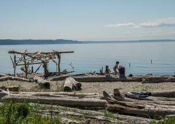 a group of kids sit on a log on the beach next to a driftwood arch