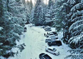 A forest road leading to a parking area that comfortably fits 12 vehicles , both covered in snow, surrounded by snow-dusted trees on both sides. 